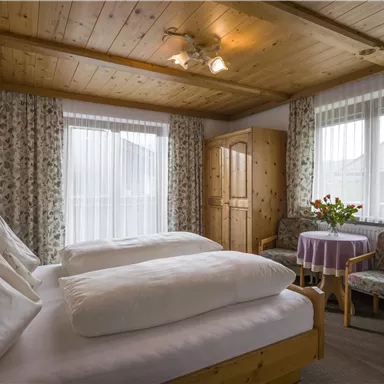 A cozy bedroom with two beds and a wooden ceiling. Daylight streams through the windows, and a table with flowers creates a pleasant atmosphere.