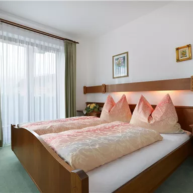 A cozy bedroom with a double bed and two decorative pillows. Bright curtains let in a lot of daylight, and some pictures are hanging on the wall.