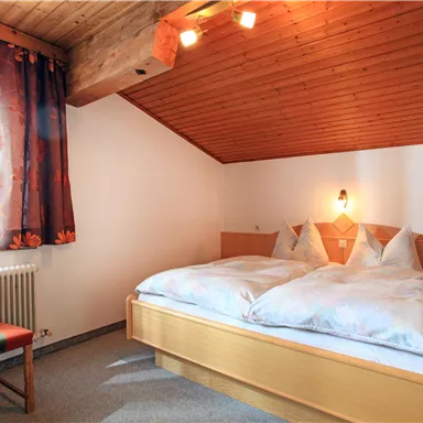 A cozy bedroom with a double bed and a chair. The walls are clad in wood and the window lets in plenty of daylight.