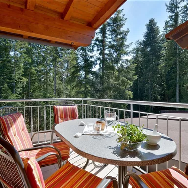 A cozy balcony with a table and two chairs, surrounded by green trees. Coffee and a small plant are on the table.
