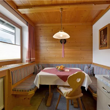 Eine gemütliche Küche mit Holzvertäfelung und einem runden Tisch. Auf dem Tisch steht eine Schale mit Obst und die Fenster sind mit Vorhängen dekoriert.