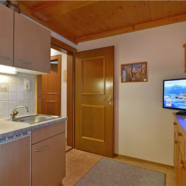 Eine gemütliche Küchenecke mit modernen Geräten und Holzverkleidung. Ein Fernseher ist ebenfalls sichtbar, dazu eine offene Tür zu einem anderen Raum.