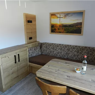 Ein gemütlicher Wohnraum mit Holzmöbeln und einer Sitzgelegenheit. An der Wand hängt ein Bild mit einer Landschaft bei Sonnenuntergang.