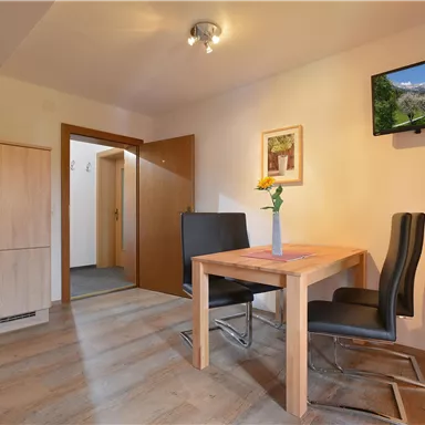 Ein heller und moderner Raum mit einem Esstisch und zwei Stühlen. An der Wand hängt ein Fernseher, und es gibt eine kleine Küche in der Ecke.