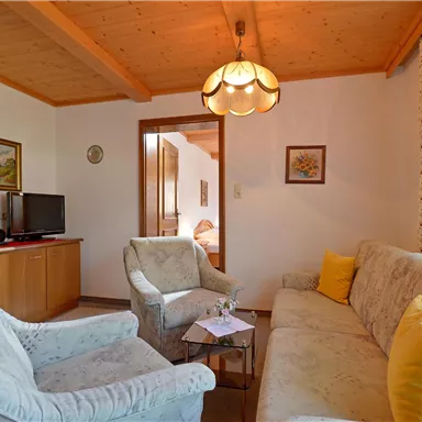 Ein gemütliches Wohnzimmer mit hellen Möbeln und einem Holzdecke. Es gibt einen Fernseher und einen kleinen Tisch in der Mitte.