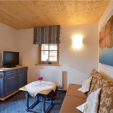 Ein gemützlicher Wohnraum mit einem Sofa und einem kleinen Tisch. An der Wand hängt ein Bild mit einem Landschaftsmotiv, und ein Fernseher steht in der Ecke.