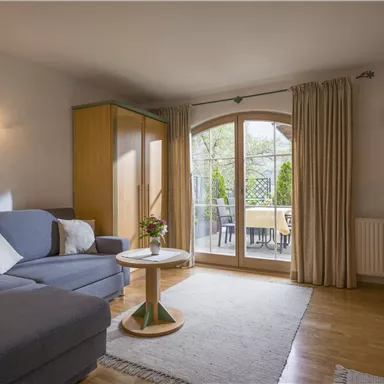 A cozy living room with a grey sofa and a small table. Large windows offer a view of the terrace and plenty of daylight.