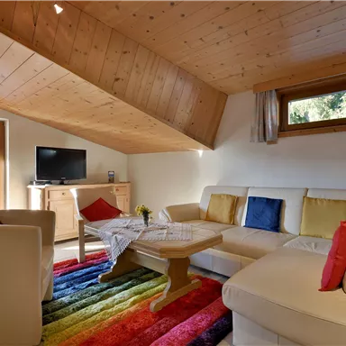 Ein gemütliches Wohnzimmer mit einem weißen Sofa und bunten Kissen. Der helle Raum hat Holzdecken und große Fenster für viel Tageslicht.