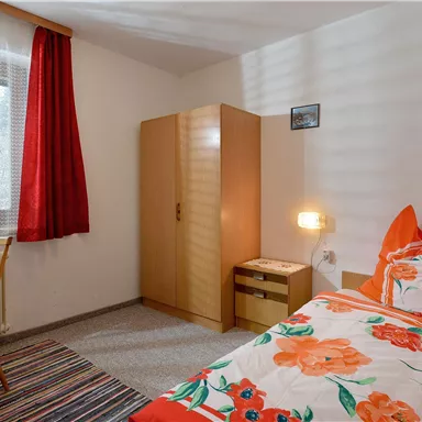 A cozy single room with a bed and colorful flower bedding. The room features a desk, a chair, and a wardrobe.