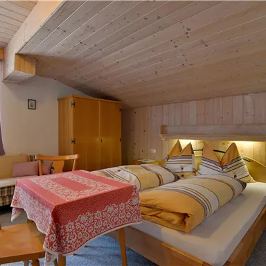 A cozy room with a double bed, wooden paneling, and soft lighting. A small table with a tablecloth and chairs completes the ambiance.