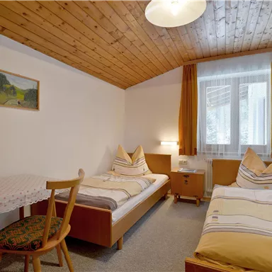 A cozy bedroom with two beds, a table, and a bright window front. The walls are in neutral colors, and there is a wooden ceiling for a warm atmosphere.