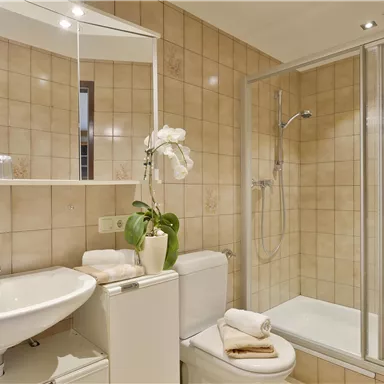 Ein modernes Badezimmer mit einer Dusche und einem Waschbecken. Die Wände sind mit hellen Fliesen dekoriert, und es gibt eine Pflanze sowie Handtücher zum Wohlfühlen.