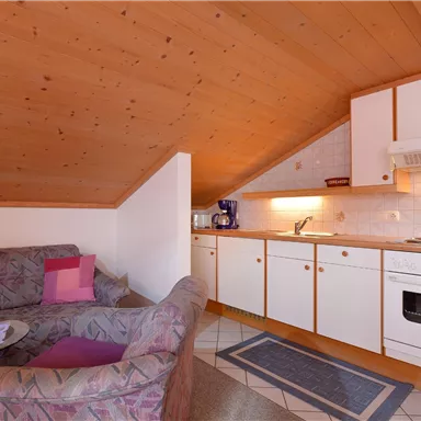 Ein gemütliches Dachzimmer mit einer Sitzecke und einer kleinen Küchenzeile. Helle Holzwände schaffen eine einladende Atmosphäre.