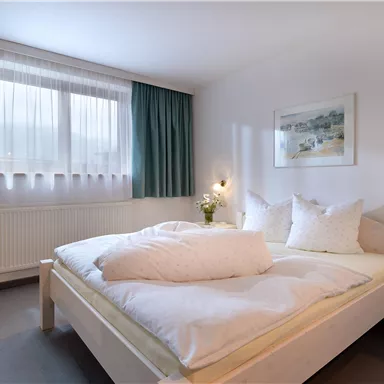 Ein helles Schlafzimmer mit einem großen Doppelbett und zwei Kissen. Die Wände sind in neutralen Farben gehalten, und es gibt Vorhänge sowie ein Fenster, das Tageslicht hereinlässt.