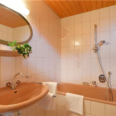 A bathroom with a bathtub and a sink. The walls are light tiled, and a round mirror hangs on the wall.
