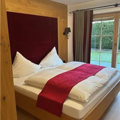 Ein gemütliches Schlafzimmer mit einem Holzrahmenbett und roten Akzenten. Große Fenster bieten Blick auf den Garten.