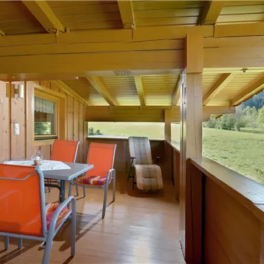 A cozy terrace with a table and chairs in orange design. The view extends over a green meadow and surrounding trees.