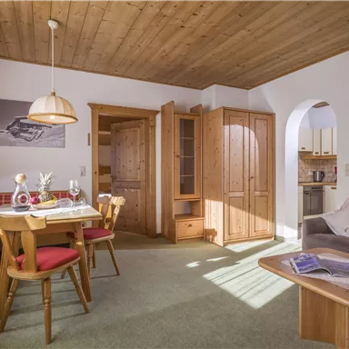 A cozy living space with a dining table and four chairs. The decor is bright and inviting, with a wooden ceiling and a small kitchenette in the background.