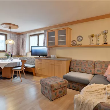 Ein gemütlicher Wohnraum mit einer Couch, einem Tisch und Stühlen. An der Wand sind mehrere Pokale ausgestellt, und das Licht ist angenehm.