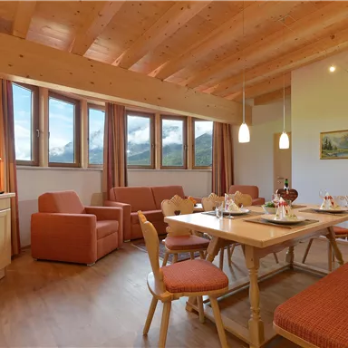Ein gemütliches Wohnzimmer mit Holzdecke und großen Fenstern. Der Raum ist mit modernen Möbeln und einem Esstisch ausgestattet.