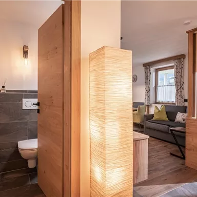 Ein modernes Badezimmer mit einer Toilette und Holzdetails. Im Hintergrund ist ein heller Wohnraum mit gemütlichen Möbeln sichtbar.