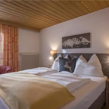 A cozy bedroom with a large bed and wooden ceiling. The atmosphere is inviting, with a red armchair and plenty of natural light.