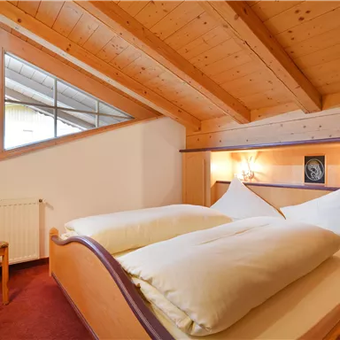 A cozy bedroom with two single beds and wooden paneling. A chair stands next to the bed, and a window lets in plenty of daylight.
