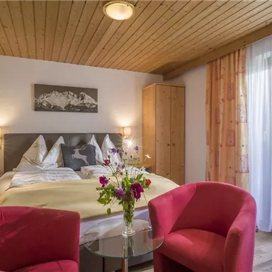 A cozy bedroom with a double bed and chairs lined with red fabric. There is also a flower vase and a window with curtains that lets in daylight.