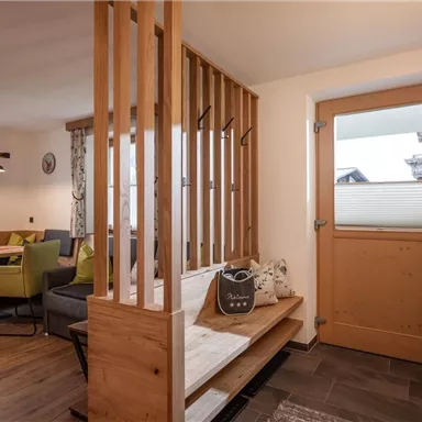 Ein einladender Eingangsbereich mit Holzverkleidung und einer gemütlichen Sitzgelegenheit. Im Hintergrund ist ein moderner Essbereich mit bunten Stühlen und einer großen Fensterfront zu sehen.