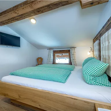 Ein gemütliches Schlafzimmer mit einem Holzrahmenbett und karierten Kissen. An der Wand hängt ein Fernseher und das Zimmer hat helle, einladende Farben.