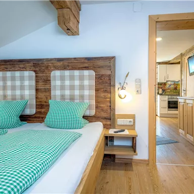 Ein gemütliches Schlafzimmer mit zwei Betten und karierten Kopfstützen. Der Raum hat Holzdetails und einen Blick auf eine Küche durch die offene Tür.