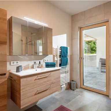 A modern bathroom with wooden cabinets and a large mirror. The door leads to an outdoor area with plenty of natural light.