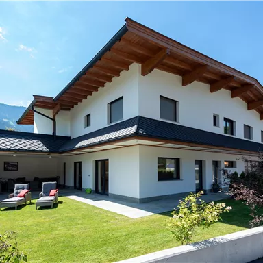 A modern house with a large garden and a terrace. In the background, there are mountains and a clear sky.