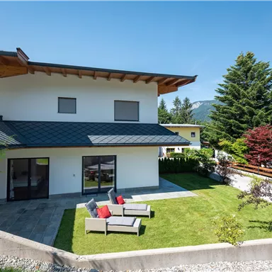 A modern house with a beautiful terrace and a well-maintained garden. In the background, mountains and trees can be seen.