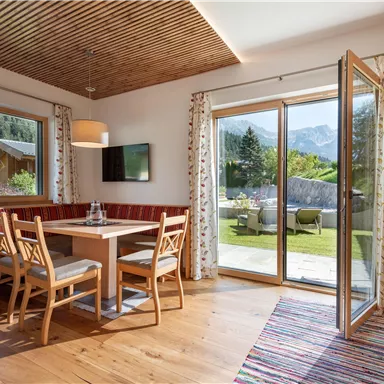 A cozy living room with a wooden ceiling, a dining table, and chairs. Through the open windows, one looks out onto a garden with a mountain view.