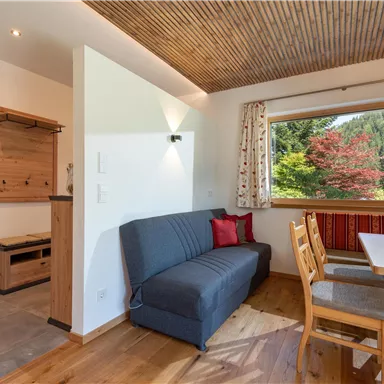 A cozy living space with wooden flooring and a modern seating area. Large windows provide a view of the green nature outside.
