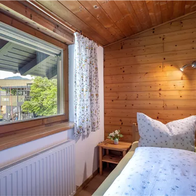 A cozy bedroom with wooden paneling and a window overlooking a green view. The beds are invitingly decorated with white pillows and blankets.