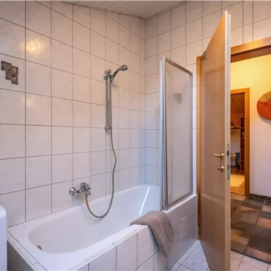 Ein Badezimmer mit einer Badewanne und einer Dusche. Die Tür ist offen und führt zu einem angrenzenden Raum.