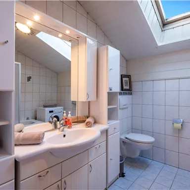 Ein helles Badezimmer mit einem Waschbecken, einem großen Spiegel und einem Fenster. Die Wände sind gefliest und es gibt Handtücher sowie Toilettenartikel.