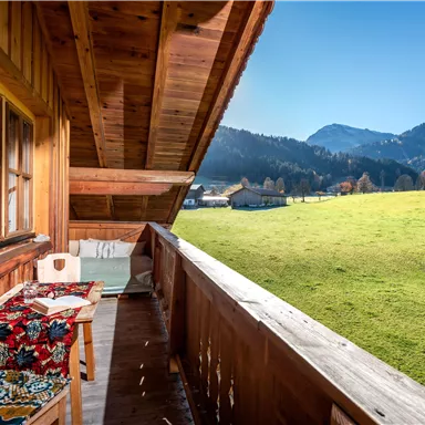 Ein gemütlicher Balkon mit Holzverkleidung und Blick auf eine grüne Wiese und Berge im Hintergrund. Der Himmel ist klar und sonnig.