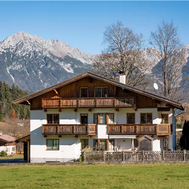 Ein schön gelegenes Haus in den Bergen mit einem großen Garten. Im Hintergrund sind majestätische Berge und blauer Himmel sichtbar.