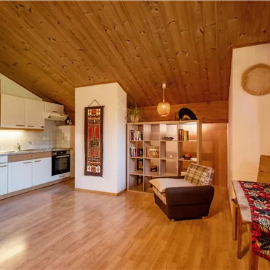 Eine gemütliche Wohnung mit Holzdecke und moderner Küche. Ein Leseecke und ein bunter Tisch sorgen für ein einladendes Ambiente.
