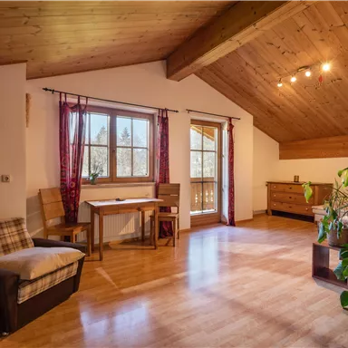 Ein helles Zimmer mit Holzdecke und großen Fenstern. Es gibt einen Tisch, Stühle und einige Pflanzen im Raum.