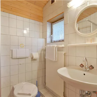 A small bathroom with a shower, a sink, and toilets. The walls are tiled with white tiles and there is a window with natural light.