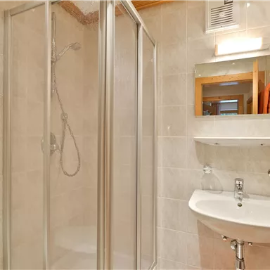 A modern bathroom with a shower cabin and a sink. The walls are light tiled and there is a mirror as well as towels nearby.