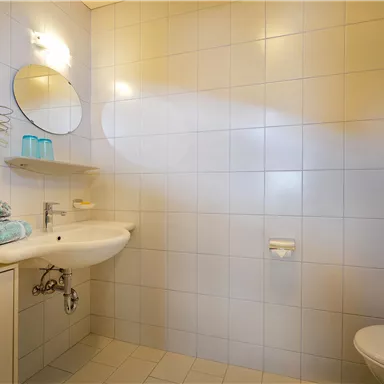 Ein modernes Badezimmer mit weißen Kacheln und einer Dusche. Auf dem Waschbecken liegen frische Handtücher und es gibt einen großen Spiegel.