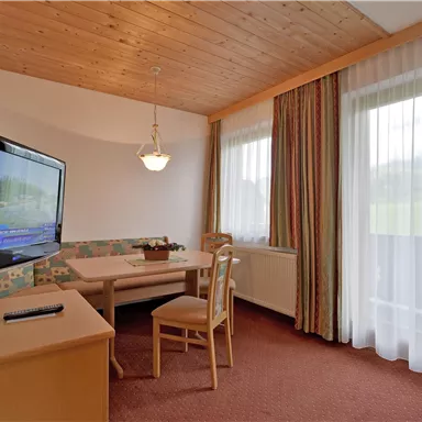 A cozy living area with a table and chairs. A television is mounted on the wall, and large windows let in plenty of light.