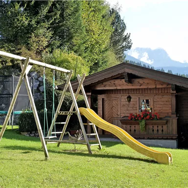 A cozy wooden house surrounded by green grass and trees. In the foreground, there is a swing and a slide.