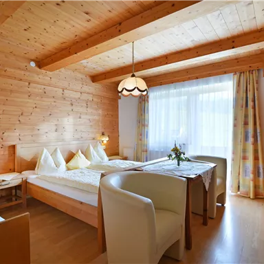Ein gemütliches Zimmer mit Holzwänden, zwei Betten und einem Tisch. Große Fenster sorgen für viel Tageslicht.