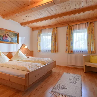 Ein gemütliches Schlafzimmer mit zwei Betten und Holzdecke. Helle Vorhänge und ein kleiner Tisch sorgen für eine einladende Atmosphäre.